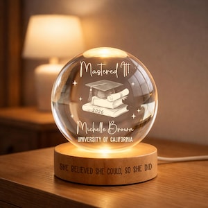 May include: A clear glass graduation sphere with the text "Mastered It!" and "Michelle Brown, University of California, 2026." The sphere sits on a wooden base with the inscription "She believed she could, so she did."