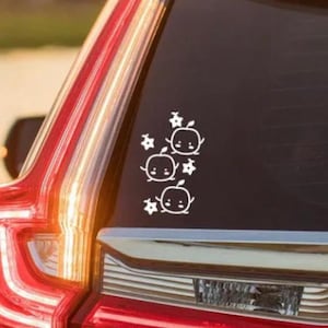 Puede incluir: Pegatina de vinilo blanco con un diseño de tres manzanas de dibujos animados con caras sonrientes y pequeñas flores. La pegatina está en la ventana trasera de un coche plateado.