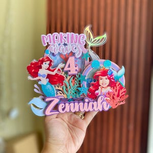 May include: A pink and purple cake topper with a mermaid theme. It features a mermaid with red hair, a number 4, and the words "Making Waves" and "Zenniah".