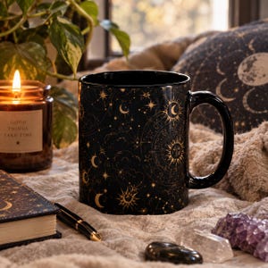 Puede incluir: Una taza de cerámica negra con un patrón celestial dorado de soles, lunas, estrellas y constelaciones. La taza tiene un acabado liso y brillante con un interior negro a juego y un asa curva, mostrando ilustraciones astronómicas.