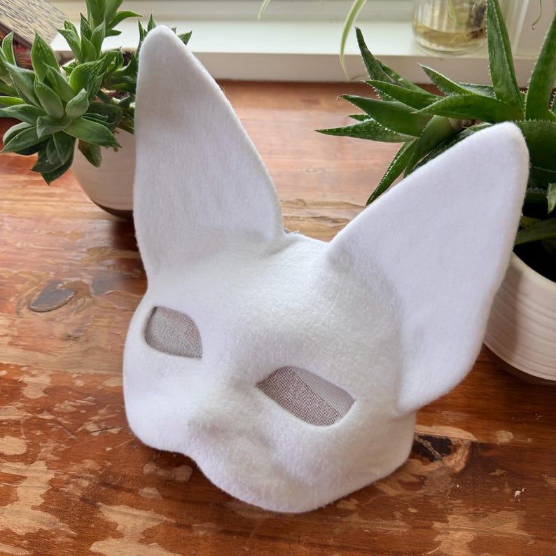 K&ouml;nnte beinhalten: Eine wei&szlig;e Katzenmaske aus Filz mit gro&szlig;en, spitzen Ohren. Die Maske hat Augen&ouml;ffnungen und ist zum Tragen bestimmt. Wahrscheinlich f&uuml;r Kost&uuml;m- oder Cosplay-Zwecke gedacht.