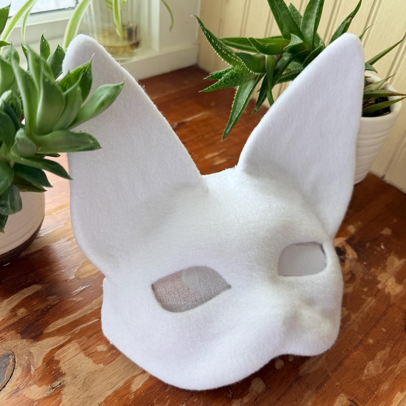 K&ouml;nnte beinhalten: Eine wei&szlig;e Katzenmaske aus Filz mit gro&szlig;en, spitzen Ohren. Die Maske hat Augen&ouml;ffnungen mit einem Netzmaterial und ist zum Tragen bestimmt. Die Maske ist ein Kost&uuml;mzubeh&ouml;r.