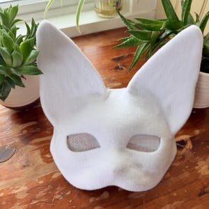 May include: A white felt mask with large, pointed ears and eye openings. The mask is designed to resemble a fox or similar animal. The eye openings are covered with a mesh material. The mask is sitting on a wooden surface.