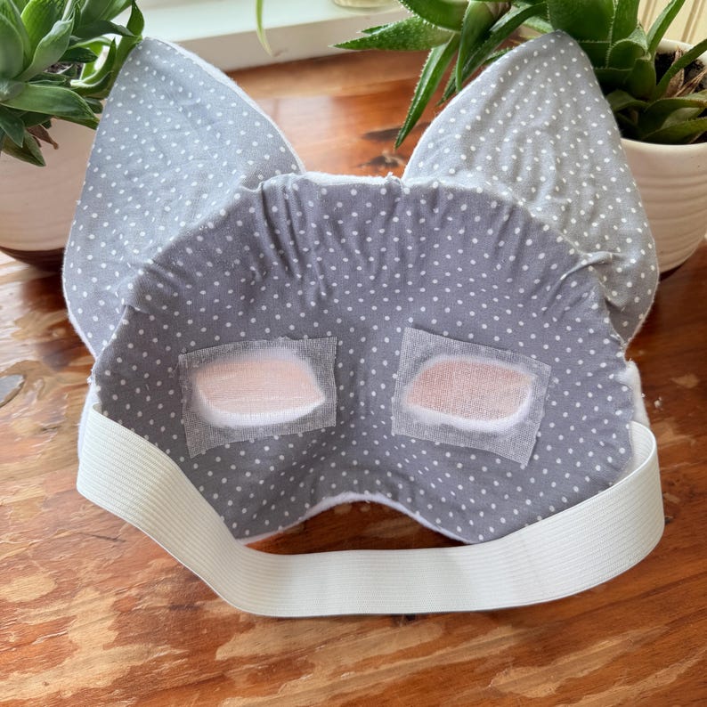 K&ouml;nnte beinhalten: Eine graue Schlafmaske mit wei&szlig;en Punkten und Katzenohren. Die Maske hat zwei rechteckige Augenabdeckungen und ein wei&szlig;es Gummiband. Entwickelt, um Licht zu blockieren und den Schlaf zu verbessern.