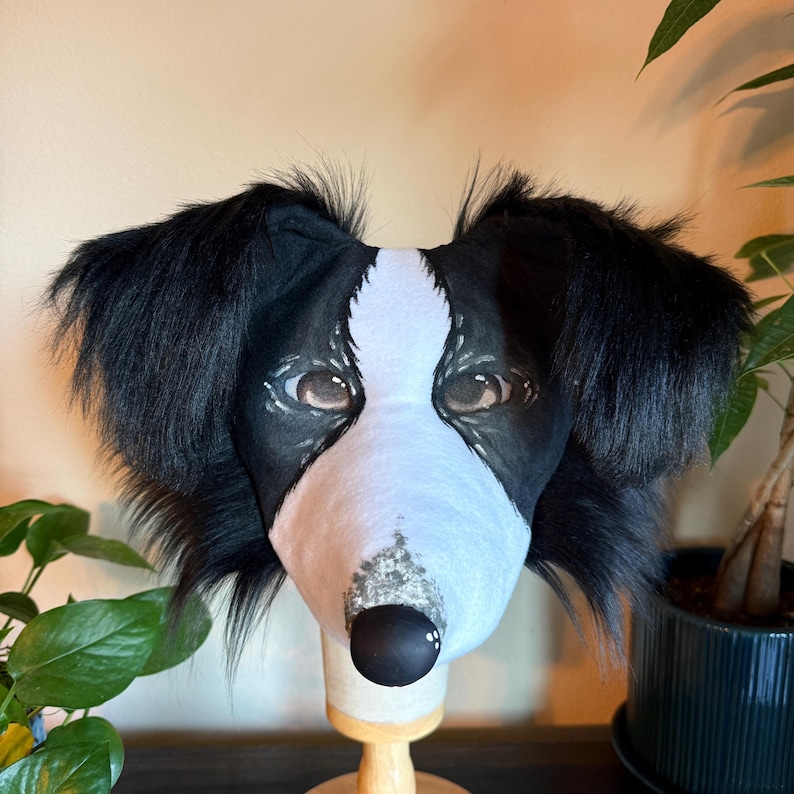 Puede incluir: Una m&aacute;scara de cabeza de perro en blanco y negro. La m&aacute;scara presenta un hocico blanco, orejas negras y ojos marrones realistas. La nariz es negra y la m&aacute;scara est&aacute; montada sobre un soporte de madera.