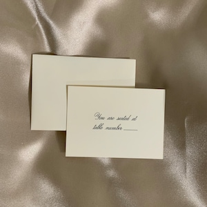 May include: A white card with black text that reads "You are seated at table number ______". The card is on a beige satin fabric.