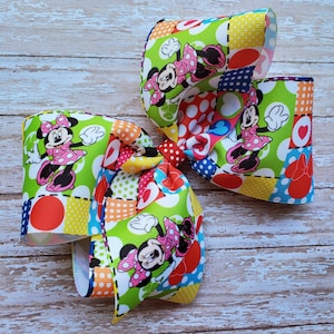 May include: A large, colorful hair bow with a patchwork design featuring Minnie Mouse. The bow is made of red, yellow, green, blue, and white fabric with polka dots and hearts.