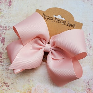 May include: A large, light pink hair bow made of grosgrain ribbon. The bow is attached to a brown tag that reads "Perfect Princess Bows" in black script. The bow has a classic, full shape.