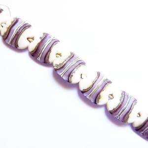 May include: A strand of white and purple glass beads with a striped pattern. The beads are oval shaped and have a glossy finish.