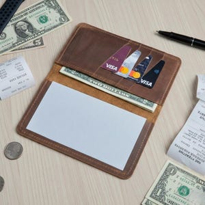 Puede incluir: Cartera de cuero marrón abierta con un bloc de notas blanco, efectivo y tarjetas de crédito. La cartera está abierta, mostrando el contenido. Se ven varios billetes y tarjetas de crédito, junto con un bolígrafo y recibos.