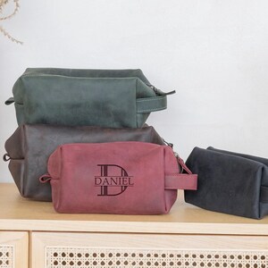 May include: A collection of leather toiletry bags in various colors: dark green, brown, burgundy, and black. The burgundy bag features the name "DANIEL" with a monogram. Each bag has a zipper closure and a handle.
