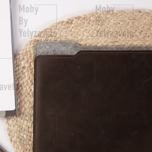 May include: A brown leather laptop sleeve with a gray felt flap and the text "Mohy By Yelyzaveta" embossed on the front. The sleeve is sitting on a natural woven mat.