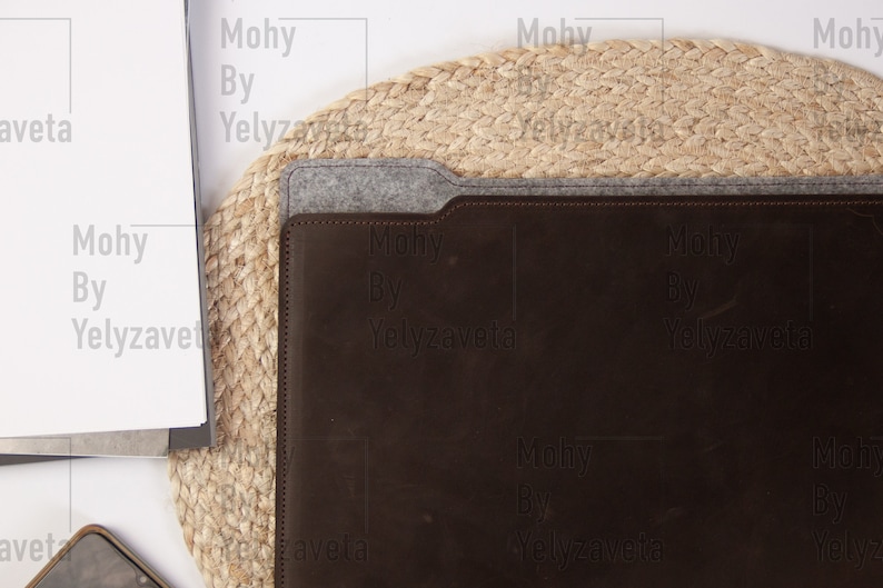 May include: A brown leather laptop sleeve with a gray felt flap and the text "Mohy By Yelyzaveta" embossed on the front. The sleeve is sitting on a natural woven mat.