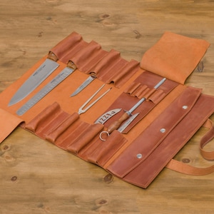 May include: An open, brown leather knife roll displaying various stainless steel knives, a honing steel, and other culinary tools. The roll is laid out on a wooden surface, showcasing the tools in individual slots.