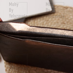 May include: A brown leather laptop sleeve with a gray felt lining. The sleeve is open and the lining is visible.