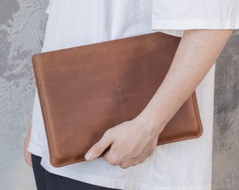 May include: A brown leather laptop sleeve with a debossed Apple logo. The sleeve is rectangular with rounded corners and a stitched border. The leather has a natural, textured appearance.
