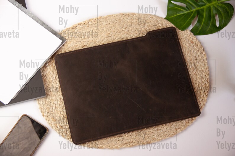 May include: A brown leather laptop sleeve on a woven jute mat. The sleeve is closed and has a subtle texture.
