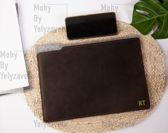 May include: A brown leather laptop sleeve with a felt lining and the initials "RT" embroidered in yellow thread. The sleeve is on a woven straw mat with a smartphone on top.