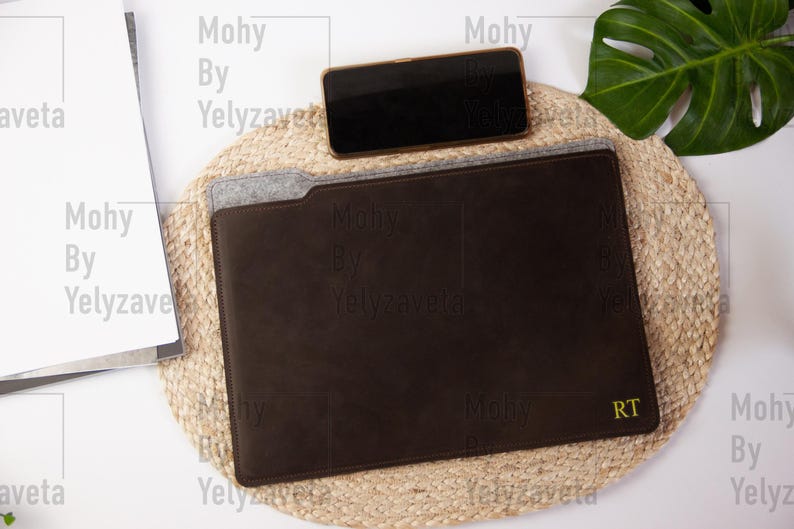 May include: A brown leather laptop sleeve with a felt lining and the initials "RT" embroidered in yellow thread. The sleeve is on a woven straw mat with a smartphone on top.