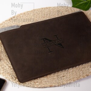 May include: A brown leather laptop sleeve with a monogrammed design. The monogram is a stylized "N" with the name "NORMAN" inscribed below it.