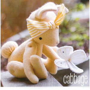 May include: A plush, cream-colored bunny rabbit toy with a yellow and white striped headband. The bunny is holding a white felt butterfly with a yellow center. The text "cabbage" is written below the bunny.