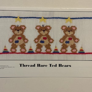 May include: A white fabric with a blue and yellow border featuring three brown teddy bears with red hearts and rainbow stacking rings. The text "Thread Bare Ted Bears" is printed below the fabric.