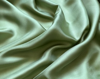 May include: Close-up of a flowing, sage green satin fabric. The fabric is draped, creating soft folds and shadows that highlight its smooth texture and sheen. The color is a muted, earthy green.