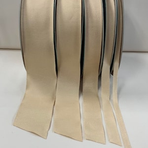 May include: Five spools of beige grosgrain ribbon, each with a different width, are unrolled and laid out on a white surface.