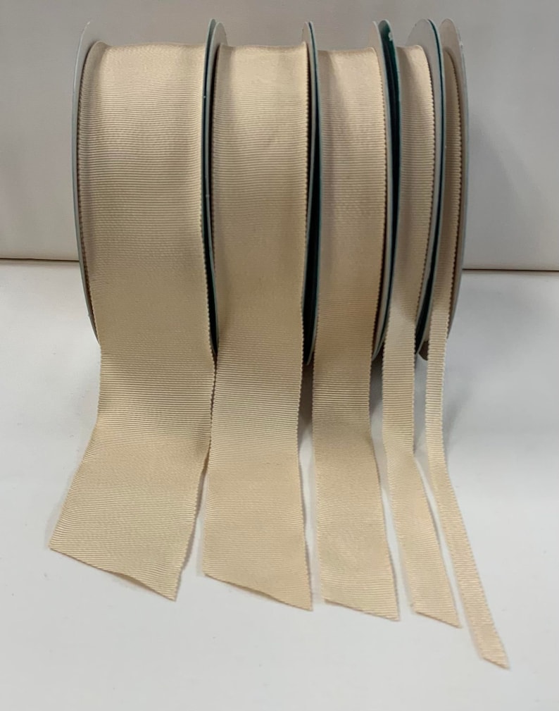 May include: Five spools of beige grosgrain ribbon, each with a different width, are unrolled and laid out on a white surface.