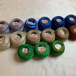May include: A collection of DMC pearl cotton thread balls in various colors, including blue, green, brown, and gray. Each ball has a label with the DMC logo and product information. The threads are tightly wound, suggesting they are new and ready for use.