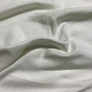 Könnte beinhalten: Nahaufnahme eines weißen, fließenden Stoffes mit weichen Falten und subtilen Schatten. Das Material scheint seidig und glatt zu sein, mit einem leichten Glanz, was auf ein luxuriöses Textil hindeutet.