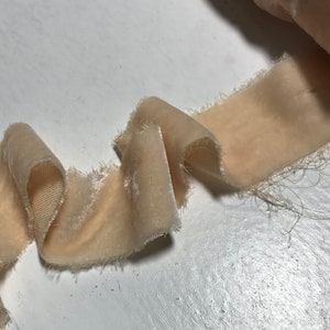 Puede incluir: Primer plano de una cinta de terciopelo de color melocotón pálido con bordes deshilachados. La cinta está doblada y retorcida, mostrando su textura y color.