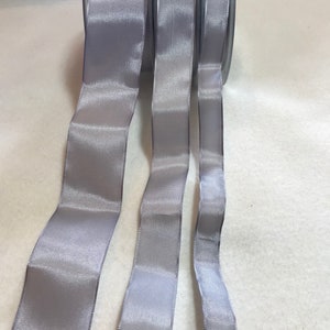 Könnte beinhalten: Drei Rollen silbernes Satinband, jede mit einer anderen Breite. Das Band ist glatt und glänzend.