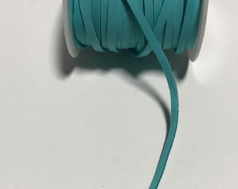 May include: A spool of teal leather cord, wound around a white plastic spool. The cord is about 1/4 inch wide and is lying on a white surface.