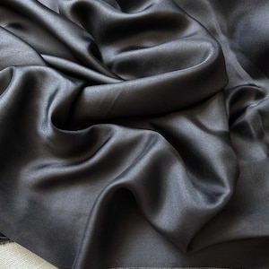 Könnte beinhalten: Nahaufnahme eines dunkelgrauen Satin-Stoffs mit glatter, fließender Textur. Der Stoff ist in weichen Falten drapiert, wodurch Schatten und Glanzlichter entstehen, die seinen Glanz betonen. Das Material wirkt luxuriös und elegant.