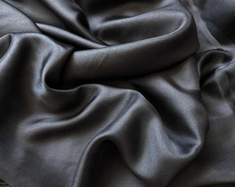 May include: Close-up of a dark gray satin fabric with a smooth, flowing texture. The fabric is draped in soft folds, creating shadows and highlights that emphasize its sheen. The material appears luxurious and elegant.