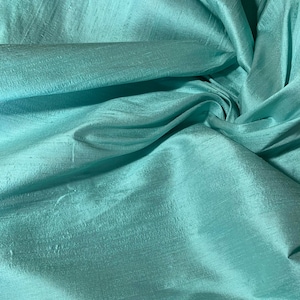 May include: A close-up of a teal blue silk fabric with a smooth, silky texture. The fabric is draped and folded, showcasing its luxurious feel.
