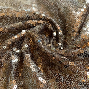 May include: Close-up of a shimmering fabric covered in small, round sequins. The sequins are primarily gold and bronze, creating a textured, reflective surface. The fabric is draped, showing folds and shadows that enhance the sparkle. Ideal for apparel and decorative projects.