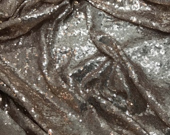 May include: Close-up of a champagne-colored fabric with a shimmering, sequined texture. The fabric is draped and folded, creating a luxurious and elegant look.