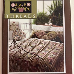 以下が含まれることがあります： Need'l Loveによる手芸本「Flower Bed Threads」。表紙にはパッチワークキルト、ボルスター、枕が置かれたベッドの写真が掲載されている。表紙のテキストには、キルト、ラグフッキング、クロスステッチ、ソーイングバードなどのプロジェクトが記載されている。