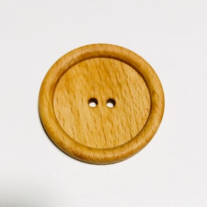 May include: A large, round wooden button with two small holes. The button is light brown with a visible wood grain and a raised outer rim. The button is likely for sewing or crafts.