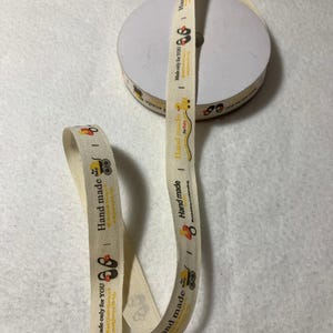 Könnte beinhalten: Eine Rolle cremefarbenes Baumwollband mit einem sich wiederholenden Muster aus einer gelben Gummiente, einem Kinderwagen und dem Text "Hand made" und "Made only for YOU".