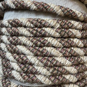 May include: Close-up of a coiled rope with a twisted design. The rope features alternating sections of beige and brown with hints of purple and green. The texture appears soft and woven, suggesting a decorative or craft material.