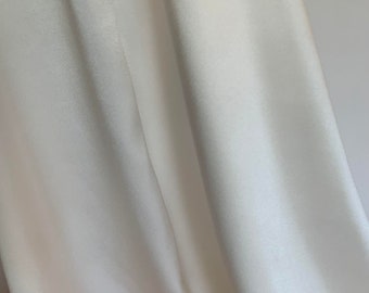 Puede incluir: Primer plano de una tela fluida de color blanco roto. El material tiene una textura suave y lisa con pliegues y reflejos sutiles, creando una sensación de movimiento y profundidad. La tela parece ligera, posiblemente sedosa.
