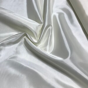 May include: A close-up of a smooth, white fabric with a silky texture. The fabric is draped and folded, creating soft, flowing lines.