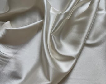 May include: A close-up of a smooth, ivory white satin fabric. The fabric is draped and folded, showcasing its luxurious texture and soft sheen.