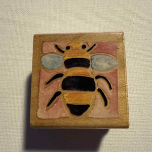 May include: A wooden block featuring a detailed illustration of a bee. The bee has black and yellow stripes, with blue wings, set against a pink background. The block has a natural wood finish.