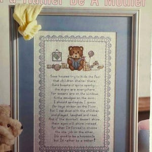 May include: Cross-stitch artwork in a silver frame with the text "I'd Rather Be A Mother." The artwork features a teddy bear reading a book, a heart, and a poem. The frame is surrounded by a light blue mat.