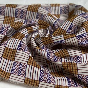 May include: Close-up of a patterned fabric with a repeating design. The fabric features a grid of squares and rectangles in shades of brown and white, with accents of dark blue and maroon. The fabric is draped and folded.
