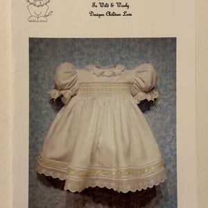 以下が含まれることがあります： 黄色い刺繍とレースのトリムが付いた白いベビー ドレス。ドレスの名前は「331 マルグリット」で、子供向けのデザインコレクションの一部です。